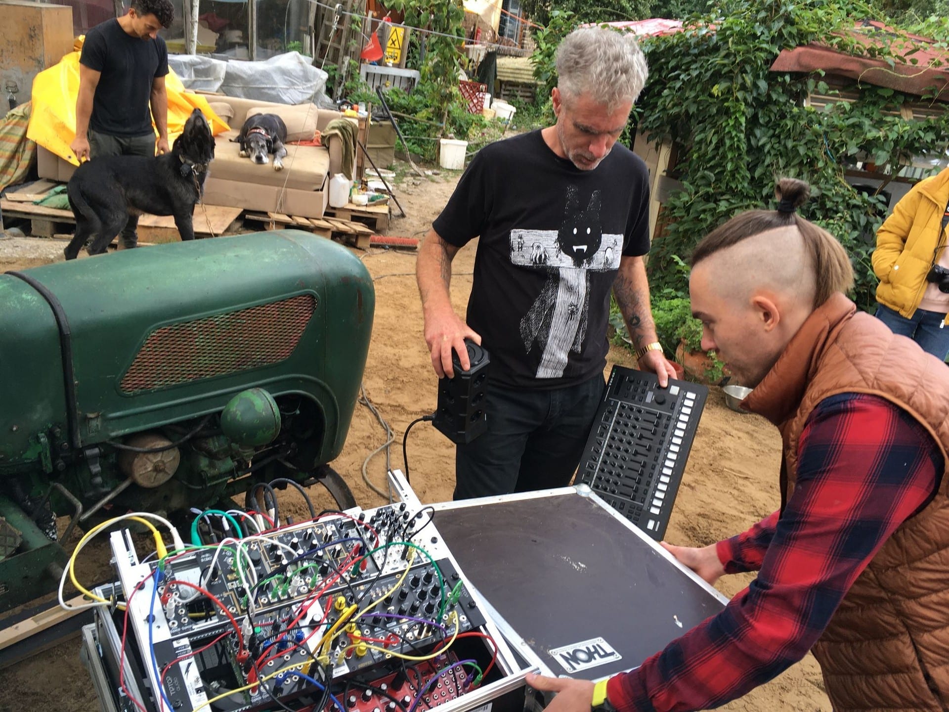 Two people examining an open modular synth case in a farmyard, with dogs on a sofa in the background