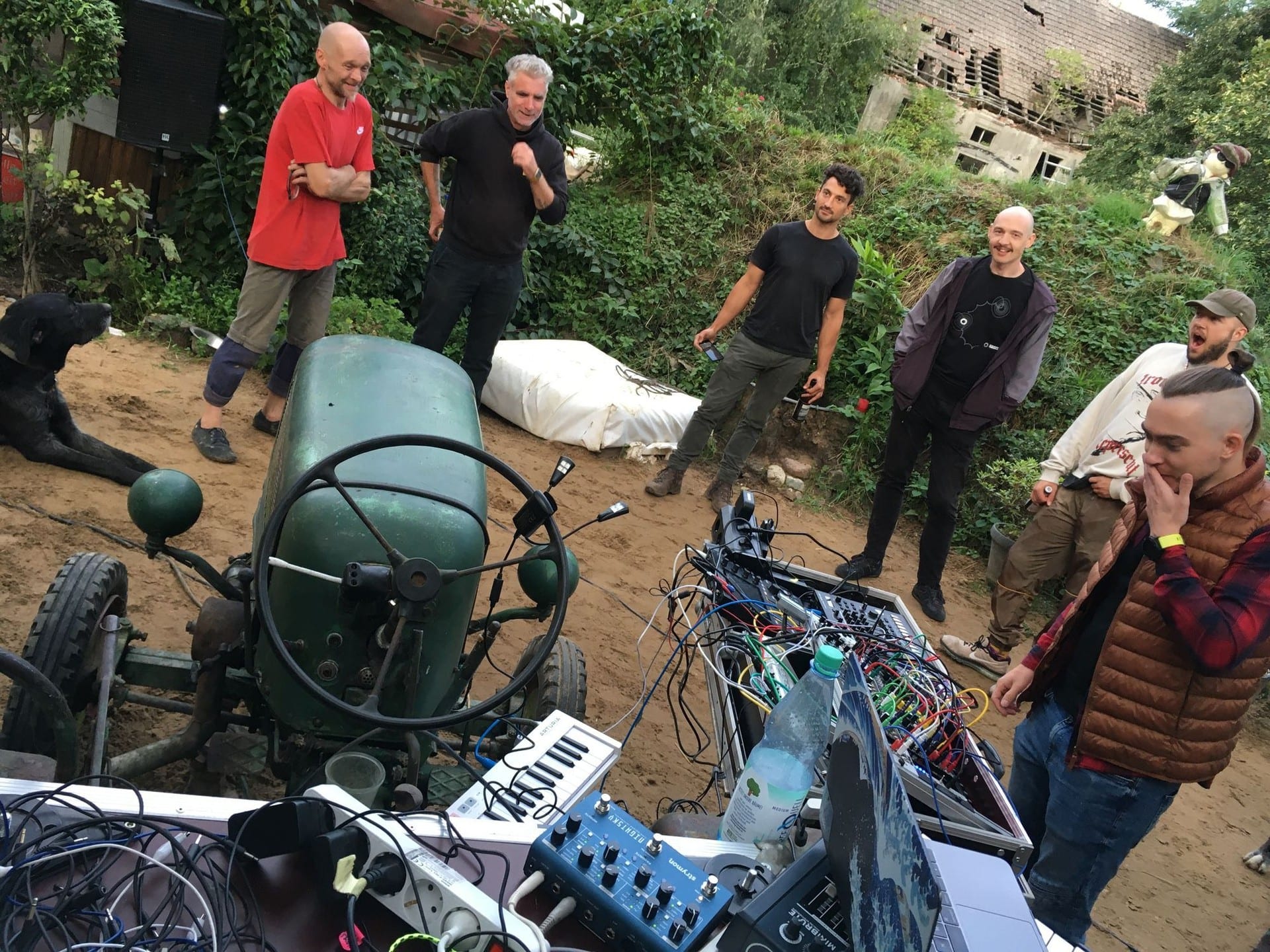 Six people standing in a sandy farmyard around a green tractor with modular gear laid out on it, a derelict brick building behind