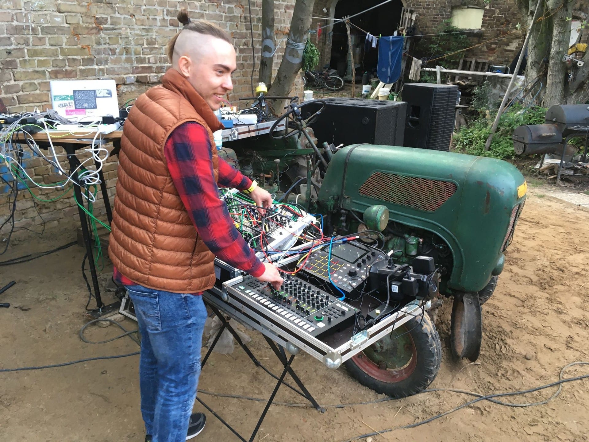 A performer in an orange gilet playing a modular rig and drum machine set up on a flight case beside the green tractor