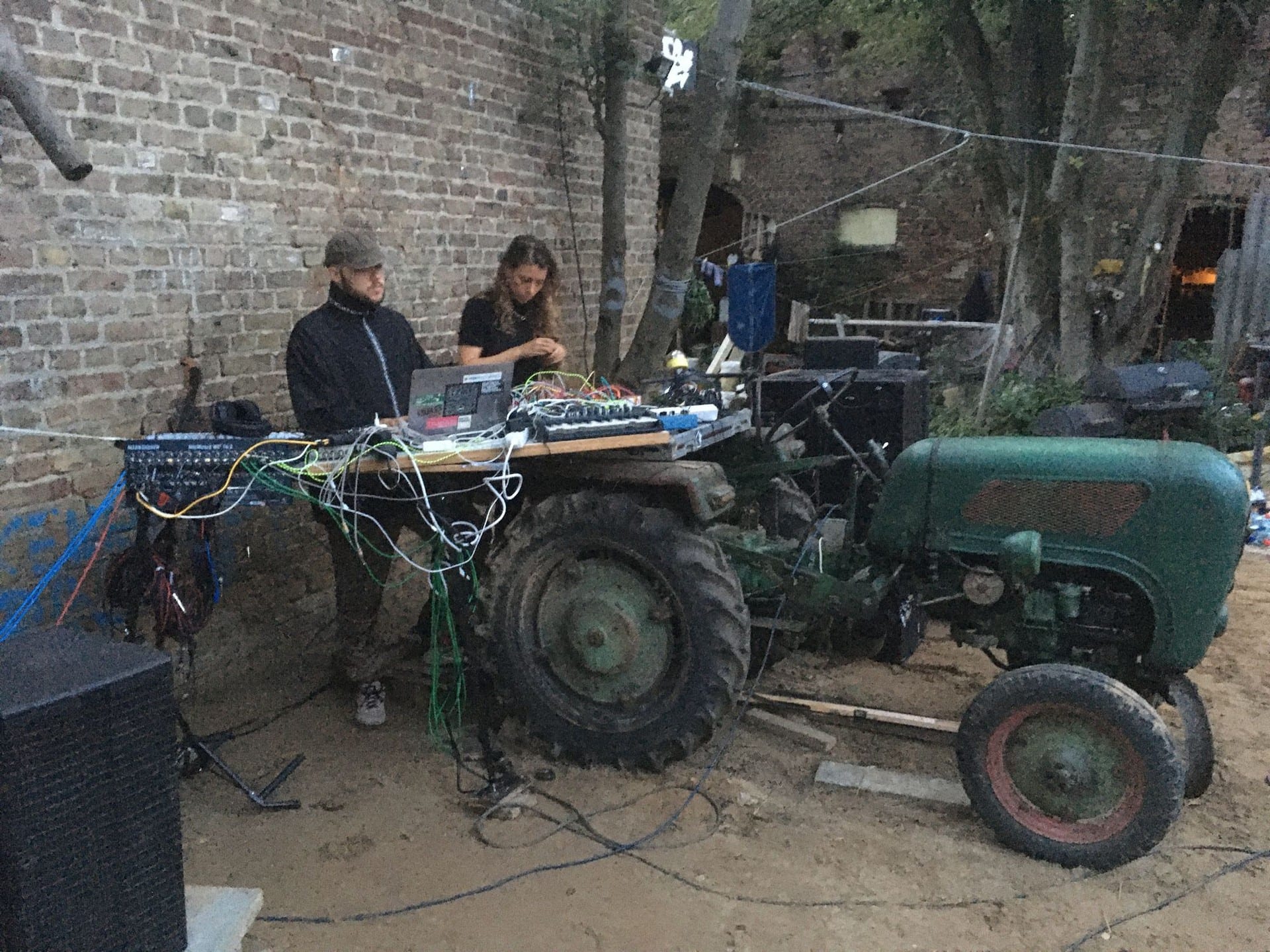 Two people performing on a laptop and modular rig balanced on a board across an old green tractor, against a brick farm wall at dusk