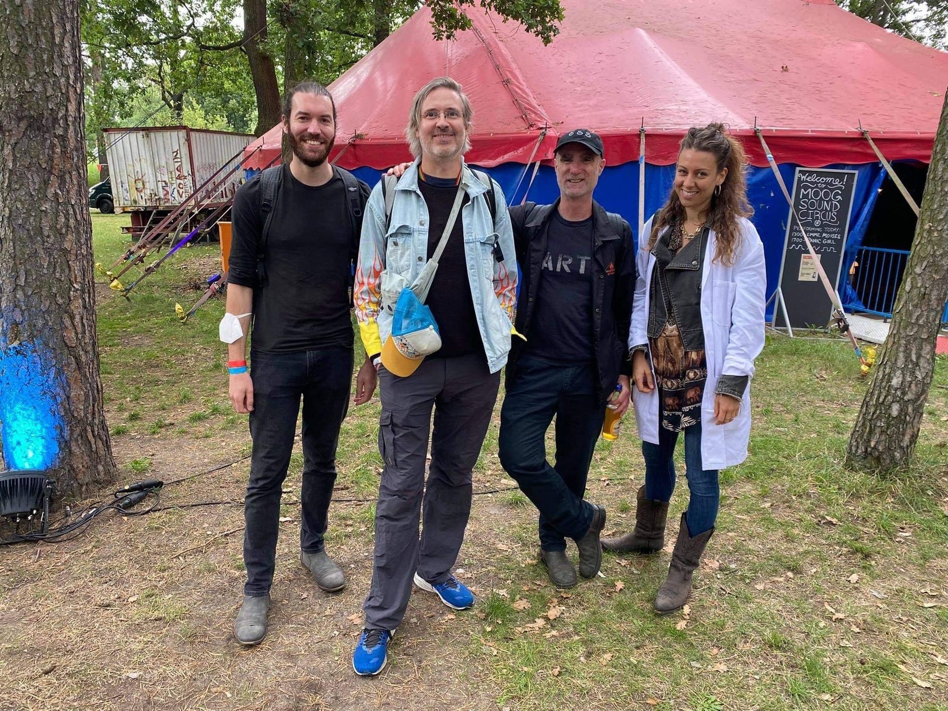 Four people standing together in front of a red and blue big-top tent at a festival, with a board reading "Welcome to Moog Sound Circus"
