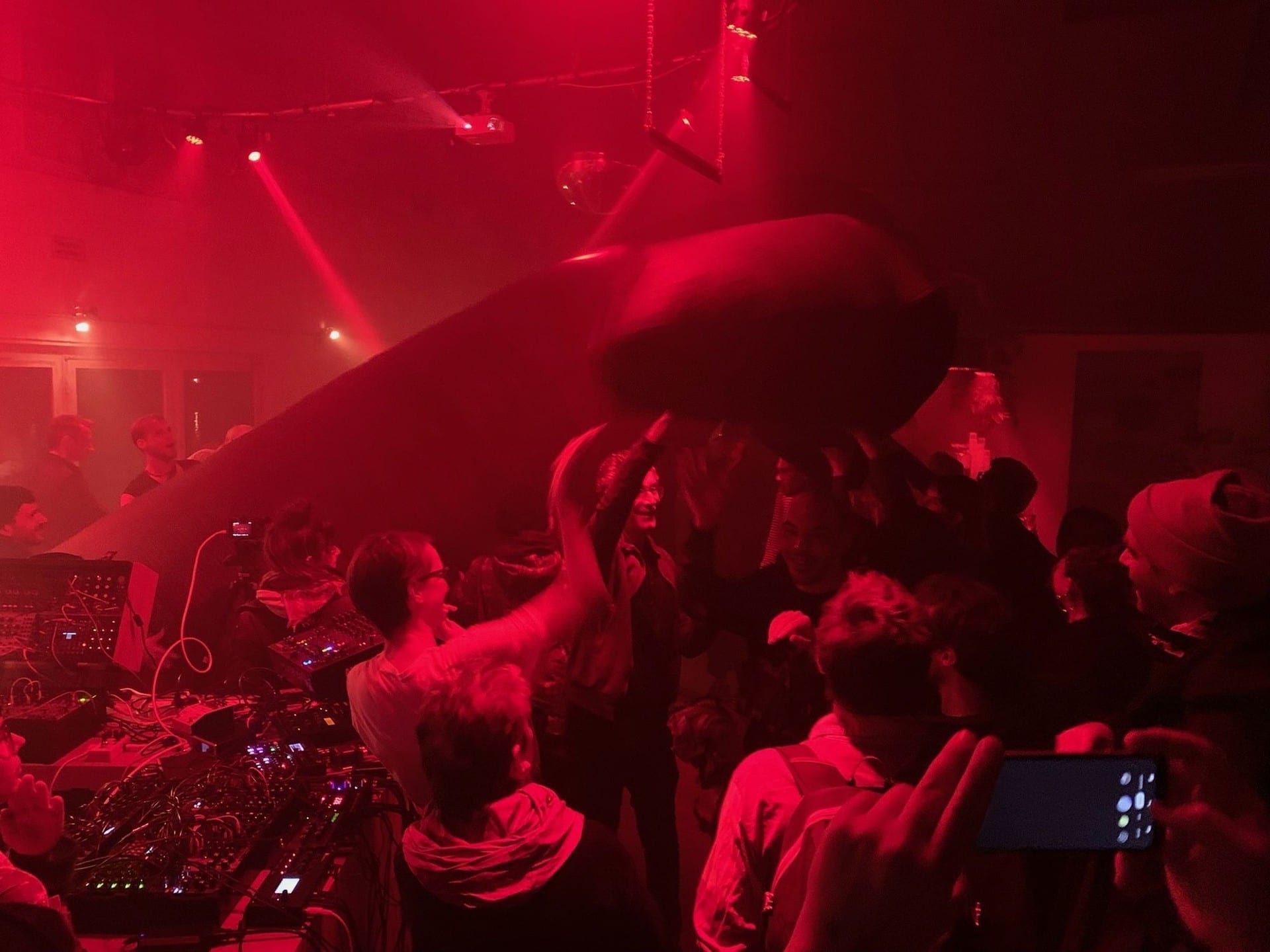A large soft sculpture held above the crowd in a red-lit club, with a modular synth rig and mixer in the foreground